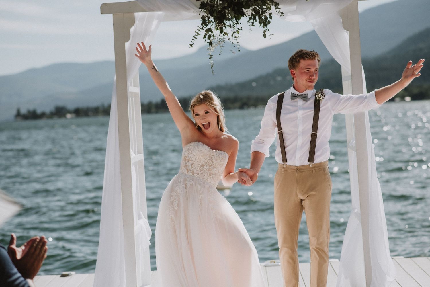 Newlyweds celebrate under white arch on waterfront dock with mountains in background.