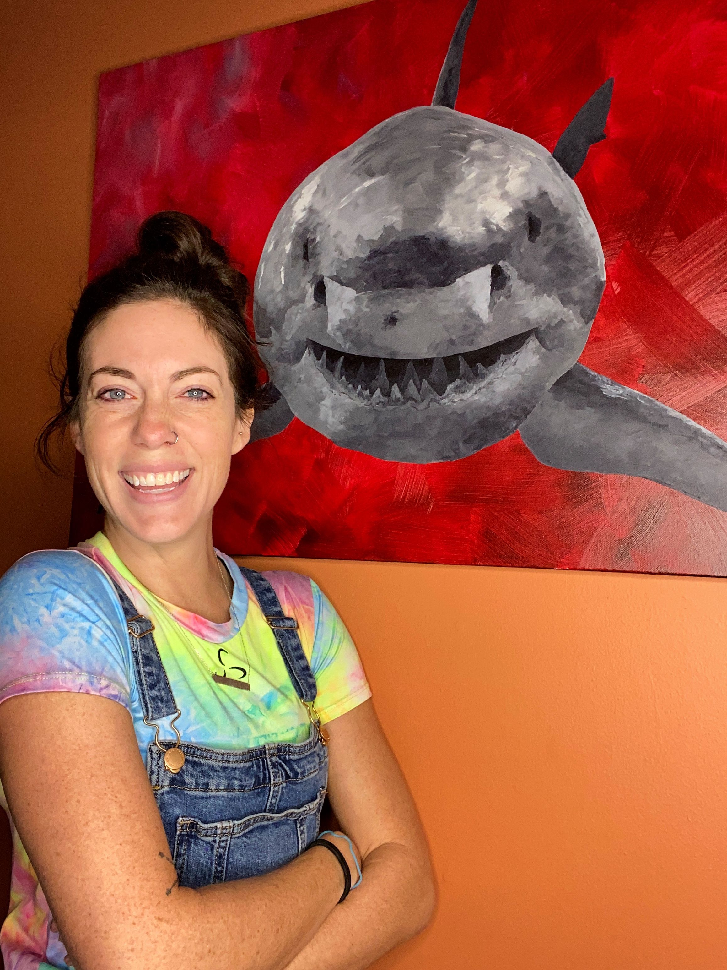 An artistic painting of a smiling grey shark on a red background displayed on a wall.