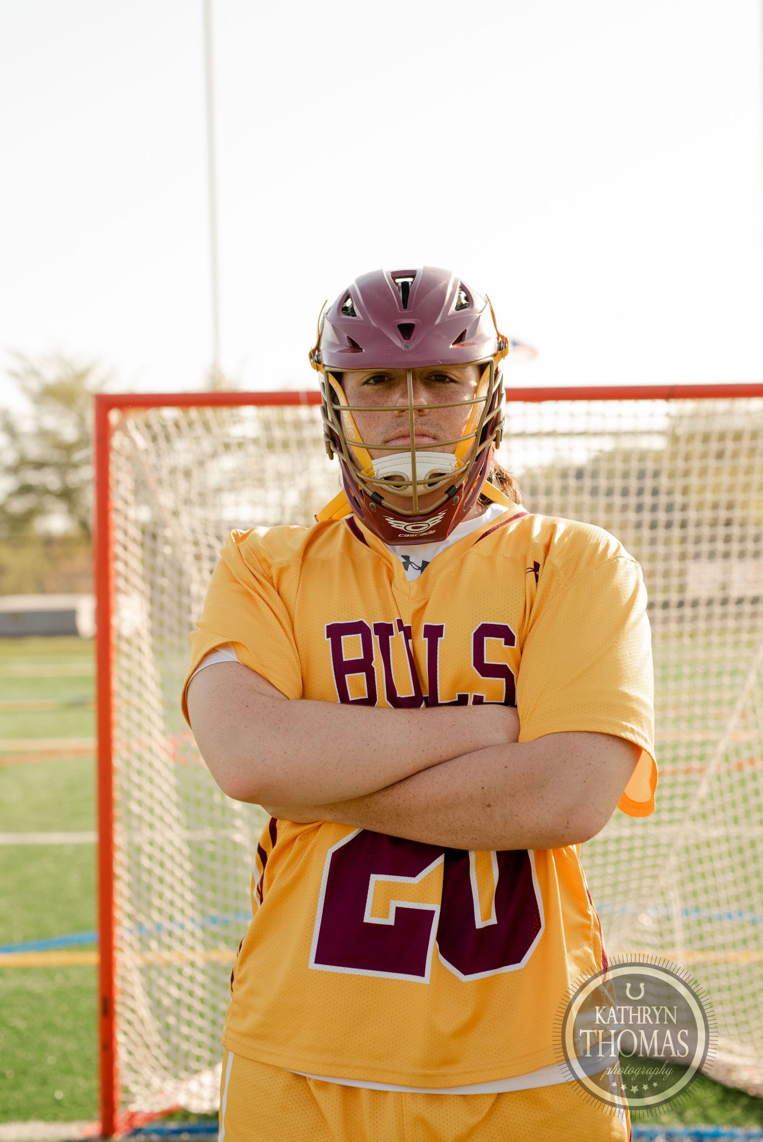 Athlete in yellow Bulls jersey number 20 with protective helmet standing by lacrosse goal.