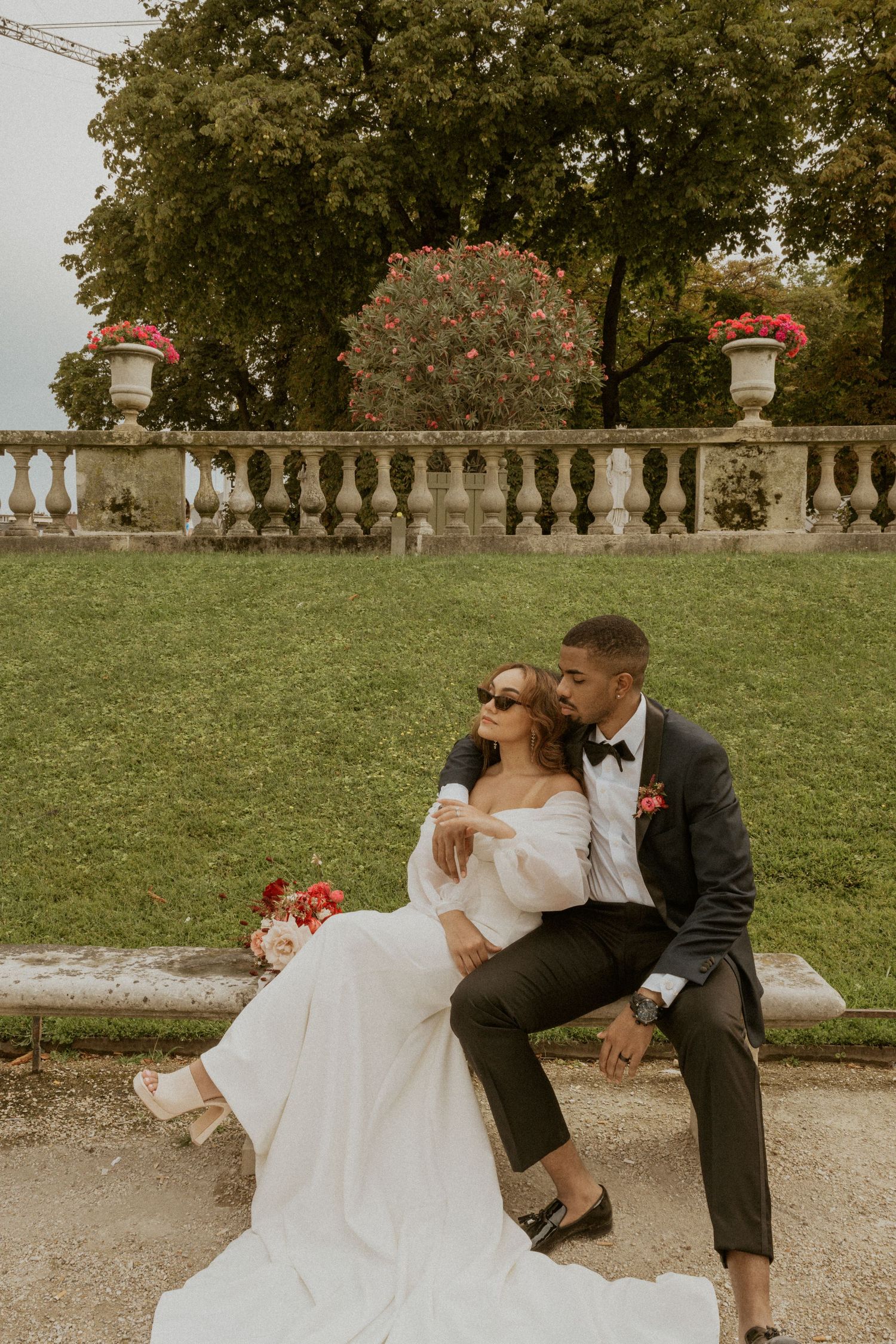 Couple in wedding attire sitting on stone balustrade with garden backdrop.