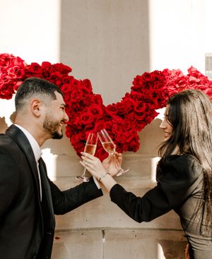Couple toasts champagne in front of large red rose heart display between columns.
