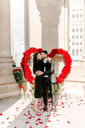Romantic couple moment in front of red rose heart display with scattered petals.