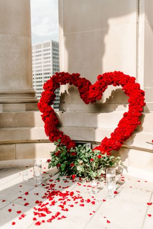 Large heart-shaped red rose display between classical columns with scattered petals.