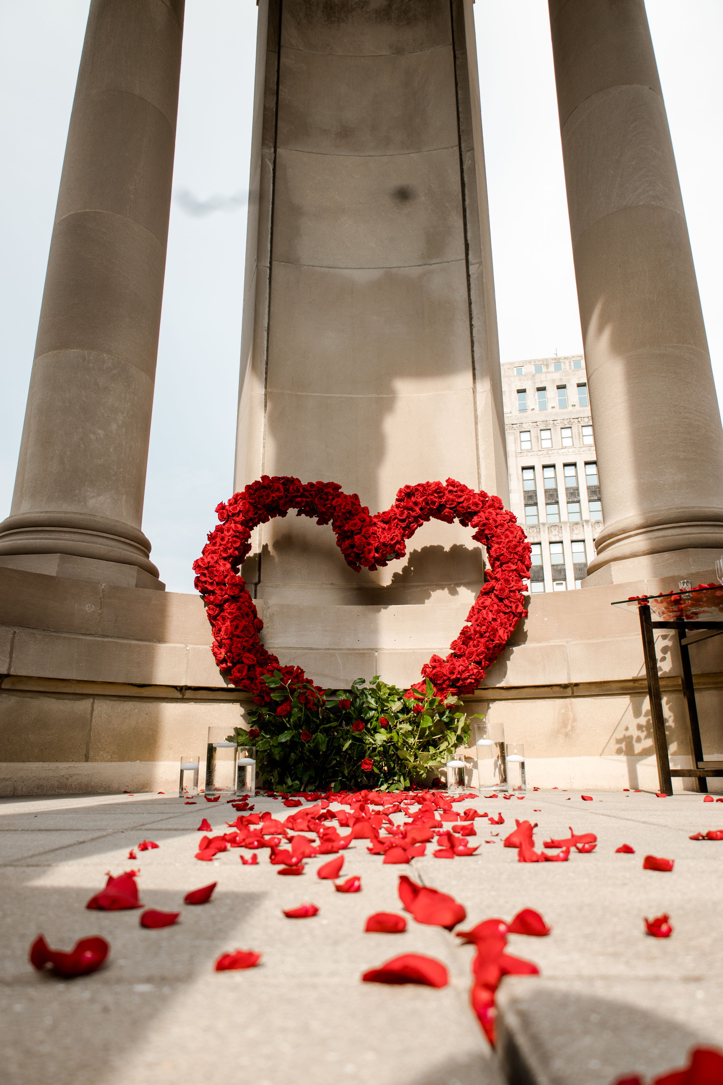 Heart-shaped red rose arrangement between elegant stone columns with scattered petals.