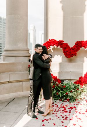 Couple embraces near red rose heart display between stone columns.