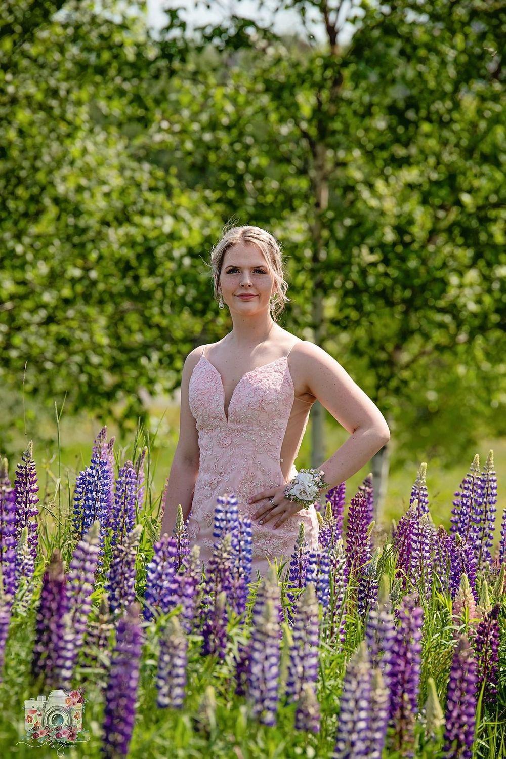A woman in a pink formal gown poses amid vibrant purple lupine flowers in a garden setting.