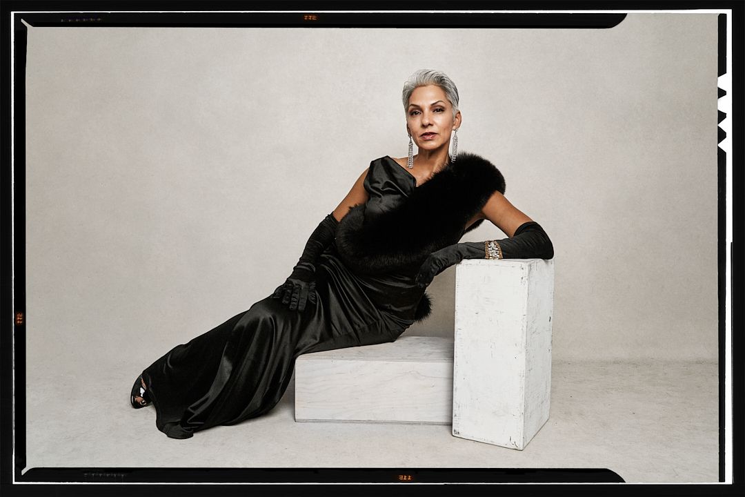 Elegant older woman in black satin gown and gloves posing on white cube in studio setting.