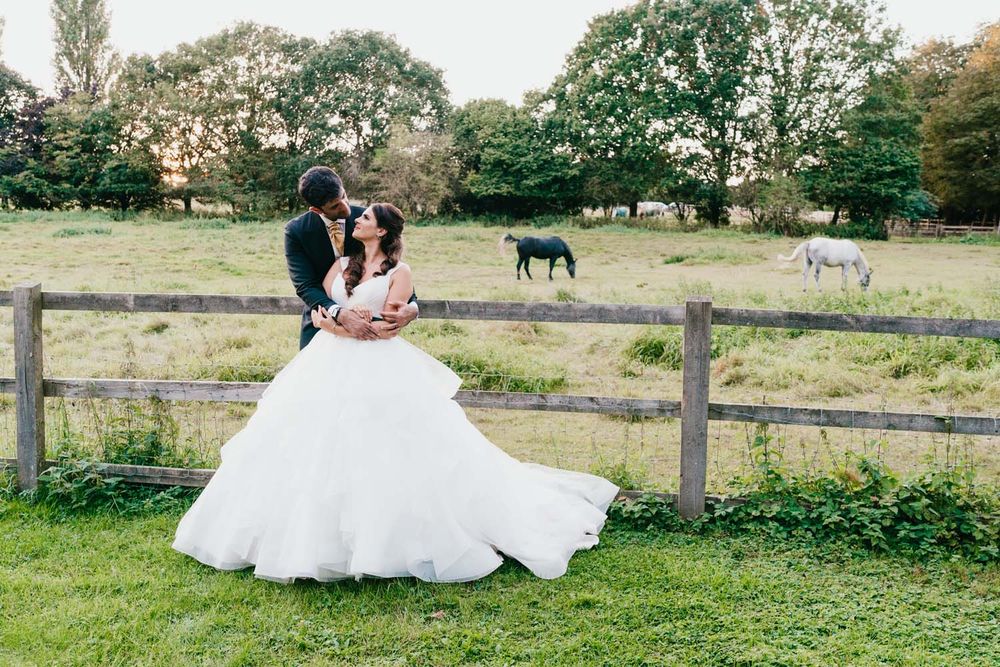 A couple shares a romantic moment by a rustic fence with horses grazing in the background.