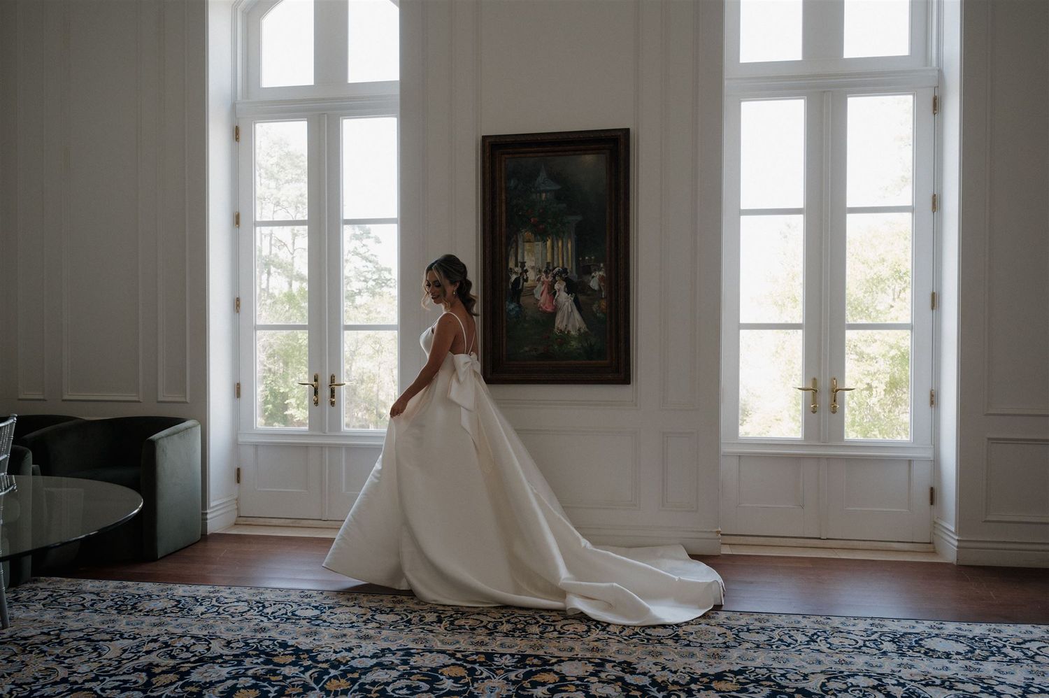 A wedding gown with a flowing train is showcased in an elegant room with tall windows and a framed painting.