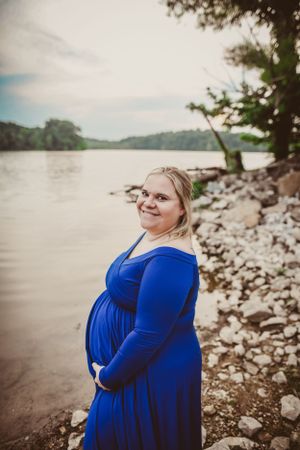 A woman in a blue dress stands by a lake shore with a joyful expression.