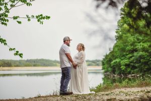 A couple stands lovingly by a serene lake surrounded by greenery.