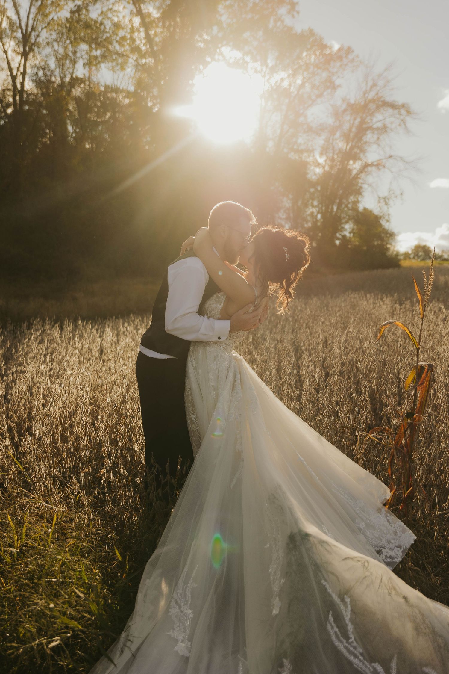 Wedding couple embracing in golden sunlit field with flowing white dress.