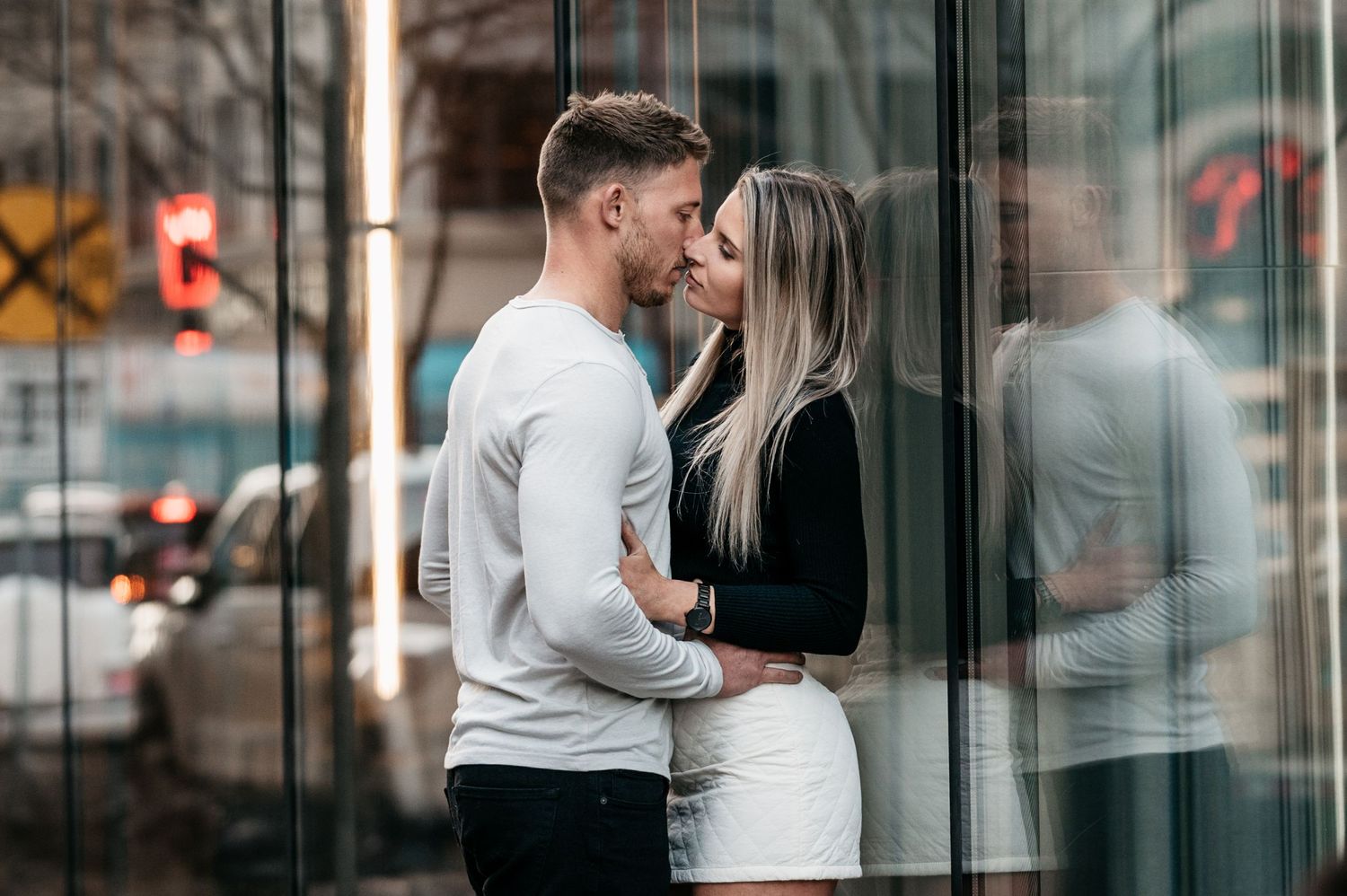Romantic couple sharing an intimate moment together in front of glass building reflections on a city street.