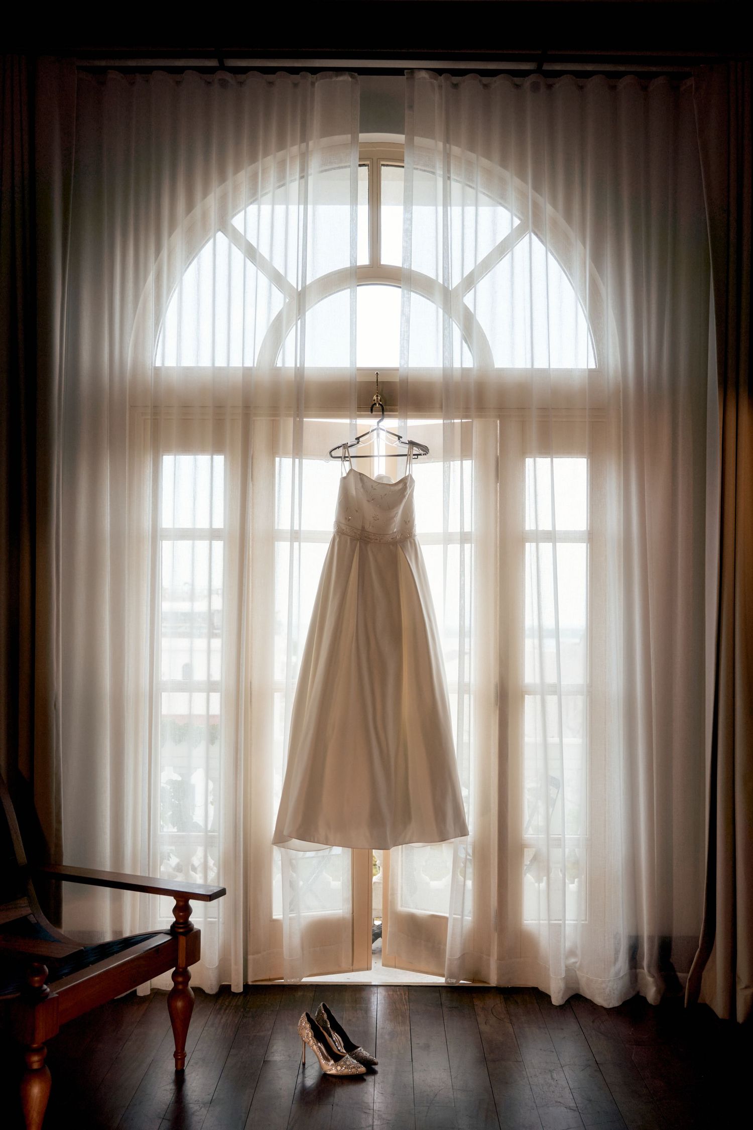 Wedding dress hanging in silhouette against arched window with sheer curtains and wooden floors.