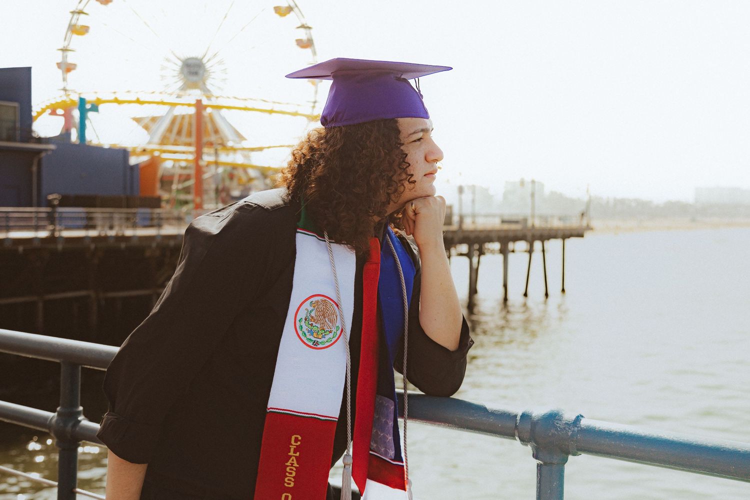 Graduate in purple cap stands thoughtfully by pier with amusement park rides visible in background.