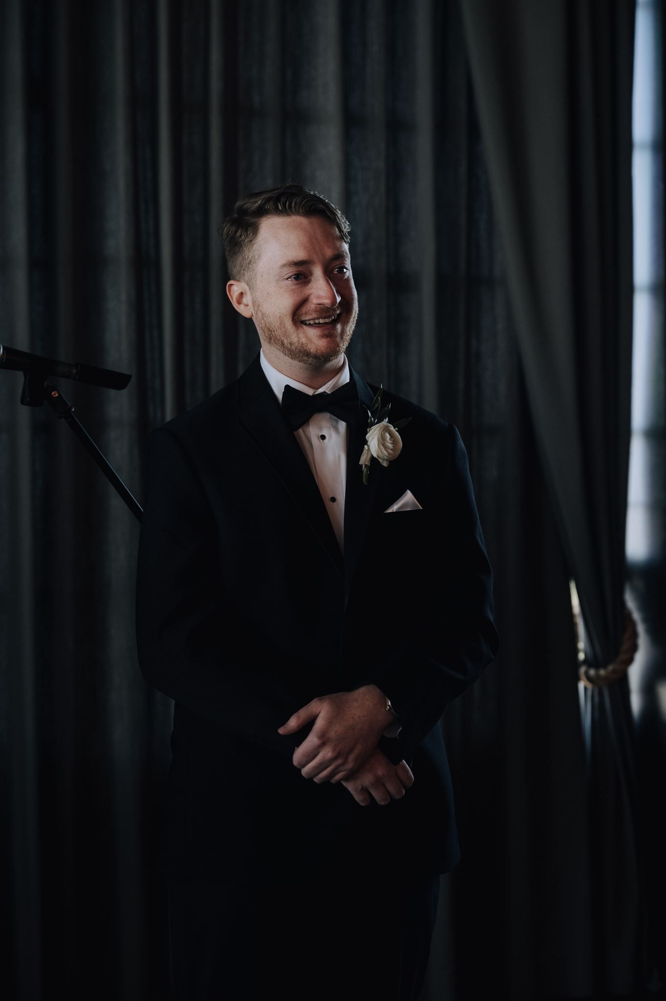 Groom in classic black tuxedo smiling against dark curtain backdrop.