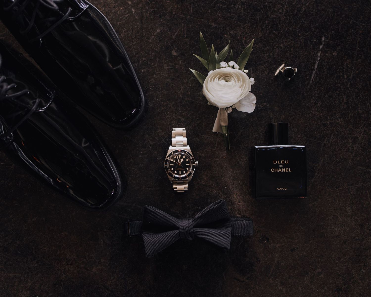Wedding accessories including black shoes, bow tie, watch and white boutonniere arranged on dark surface.