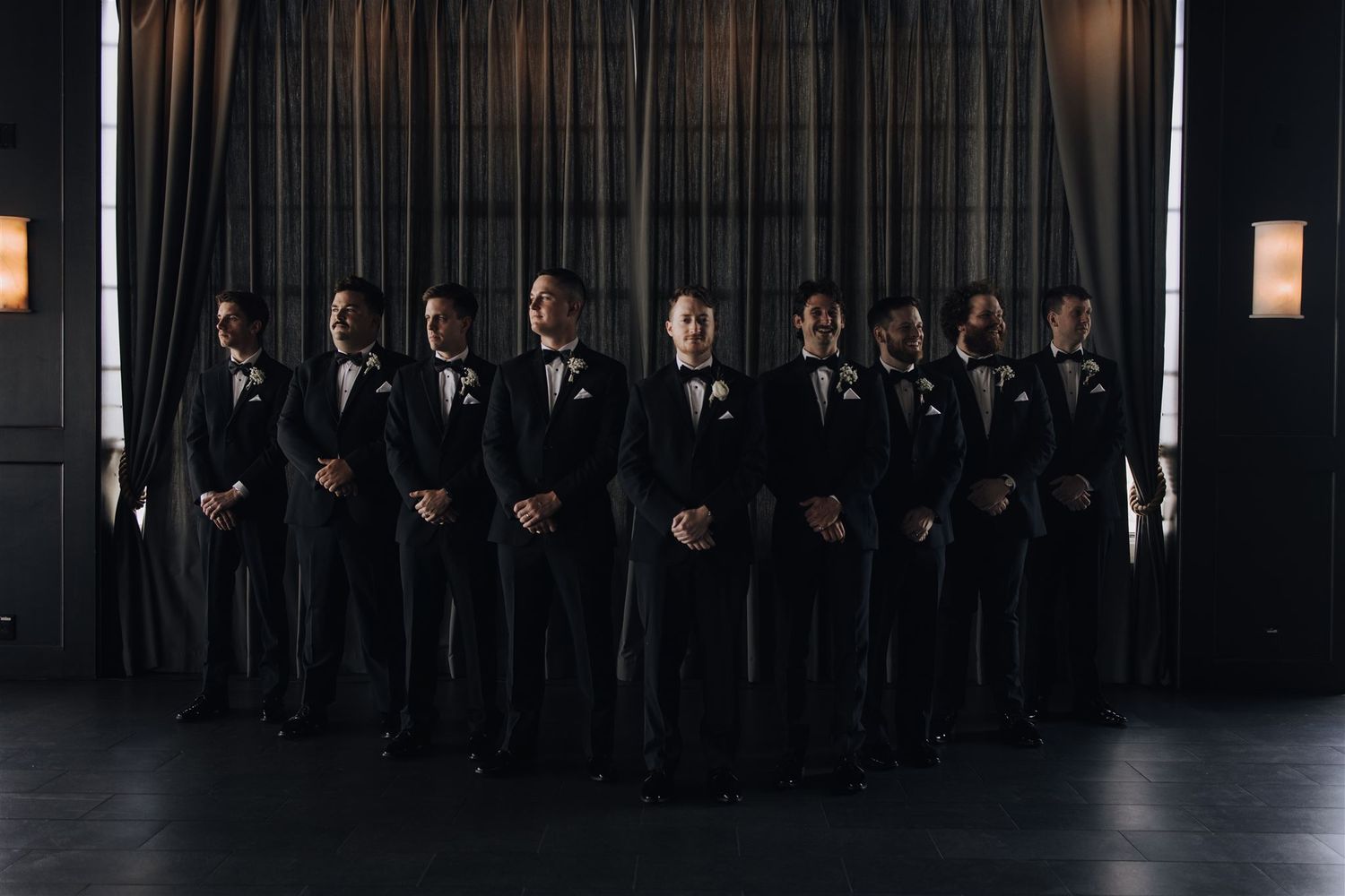 A group of groomsmen in black tuxedos pose formally against a dark curtain backdrop.