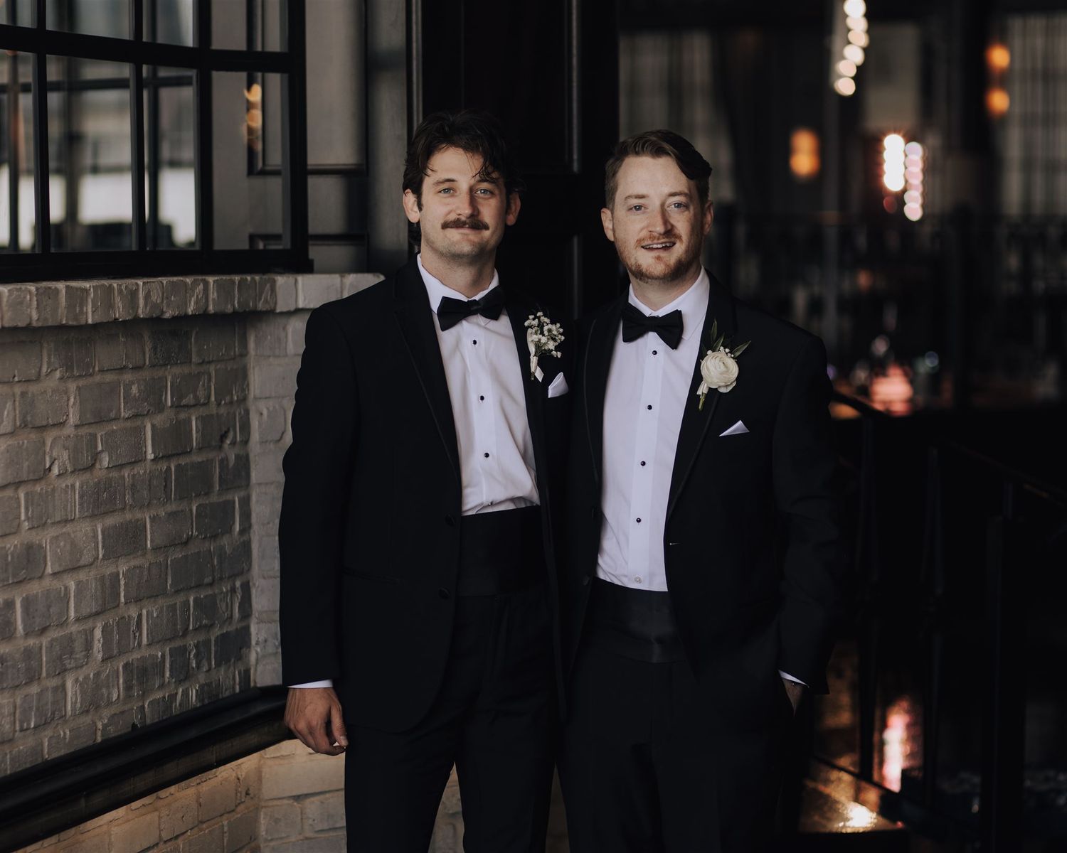 Two people in elegant black tuxedos with bow ties pose together against a brick wall at a formal event.