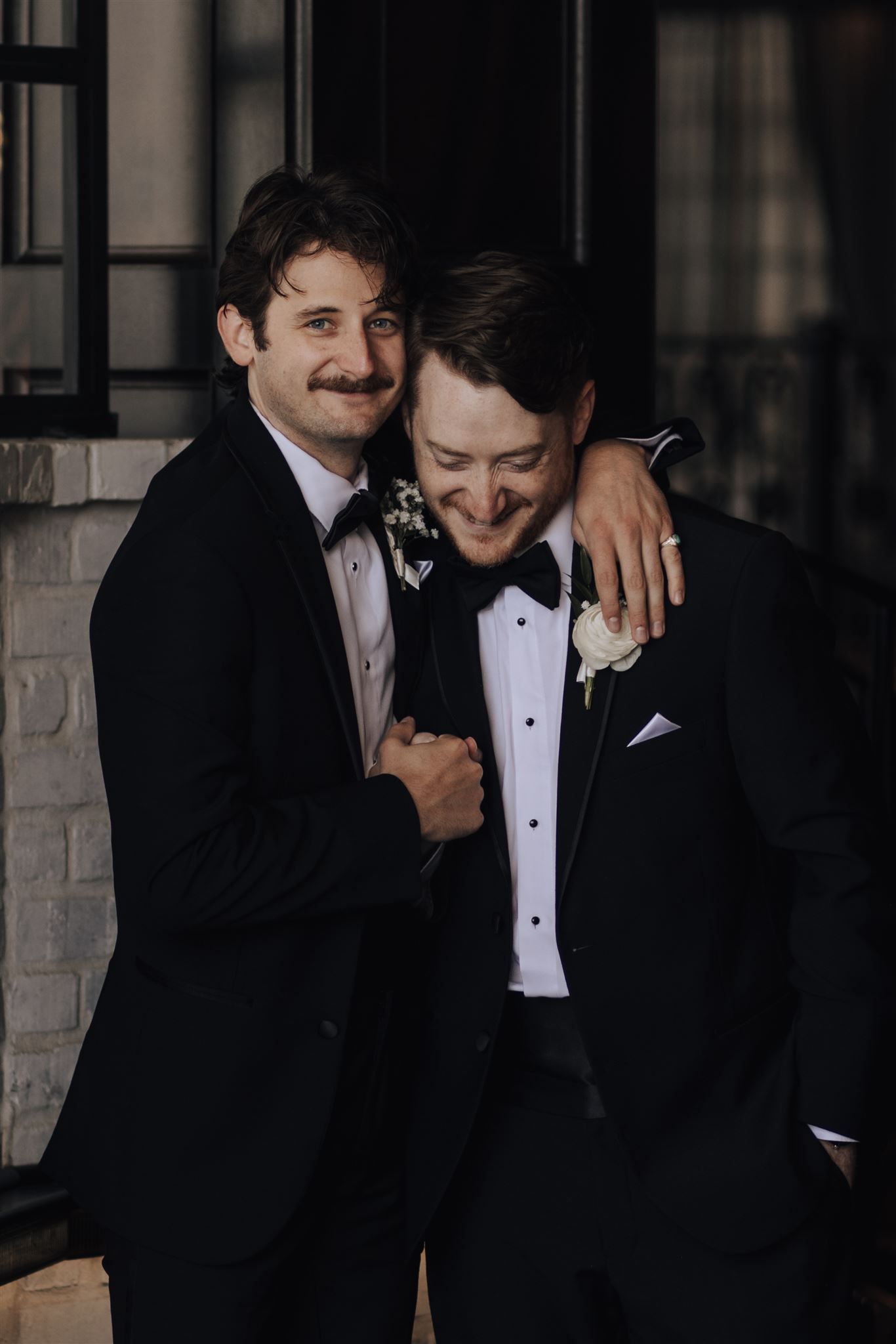 Two people share a joyful candid moment wearing tuxedos at a formal event.