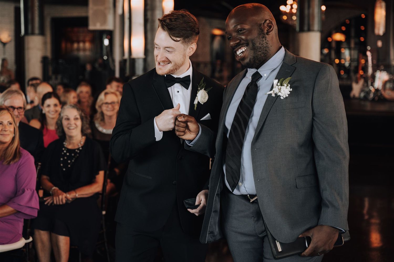 Two people sharing a joyful moment during wedding reception with guests in background.