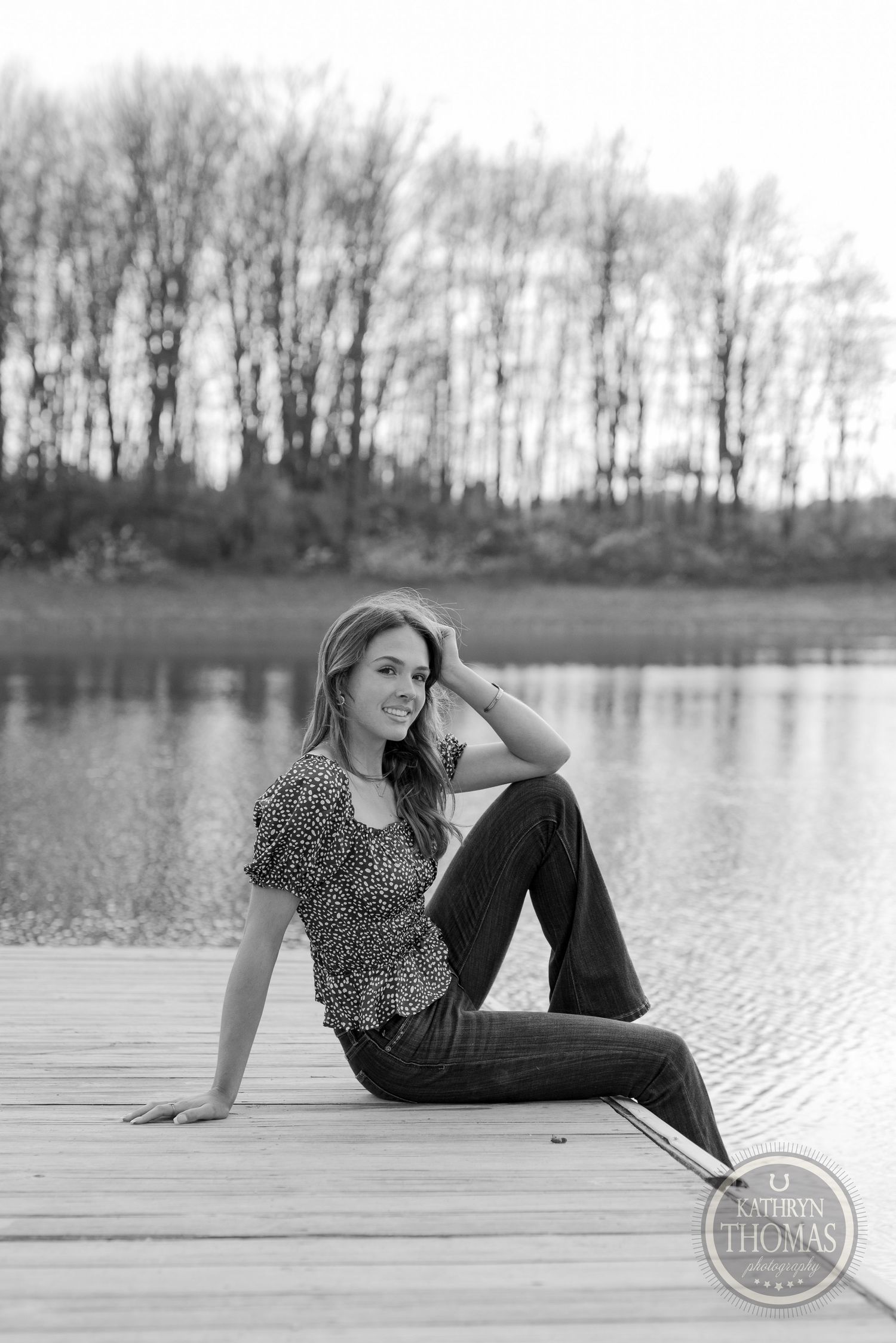 Black and white portrait of someone in casual attire sitting on a lakeside dock during autumn.