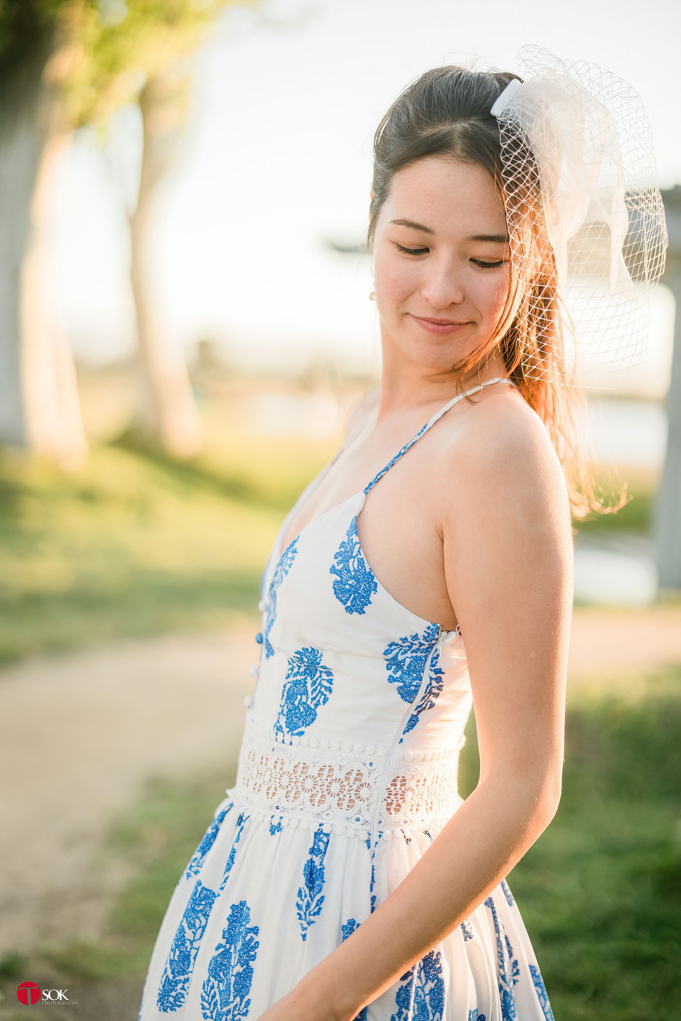 A young woman in a blue patterned dress smiles softly in warm sunlight.