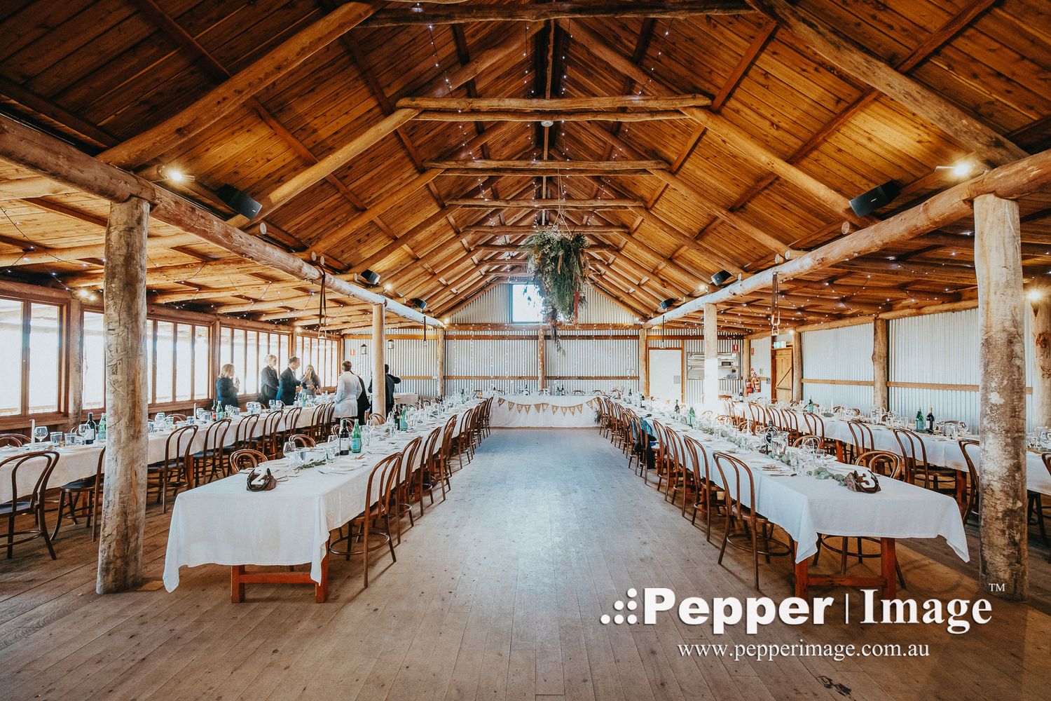 Rustic barn wedding reception venue with long white-clothed tables, wooden chairs, and warm string lights overhead.