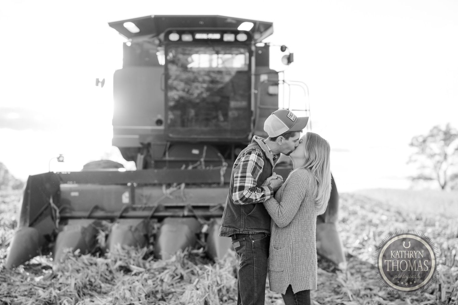 A romantic couple stands together near a combine harvester in a black and white countryside scene.