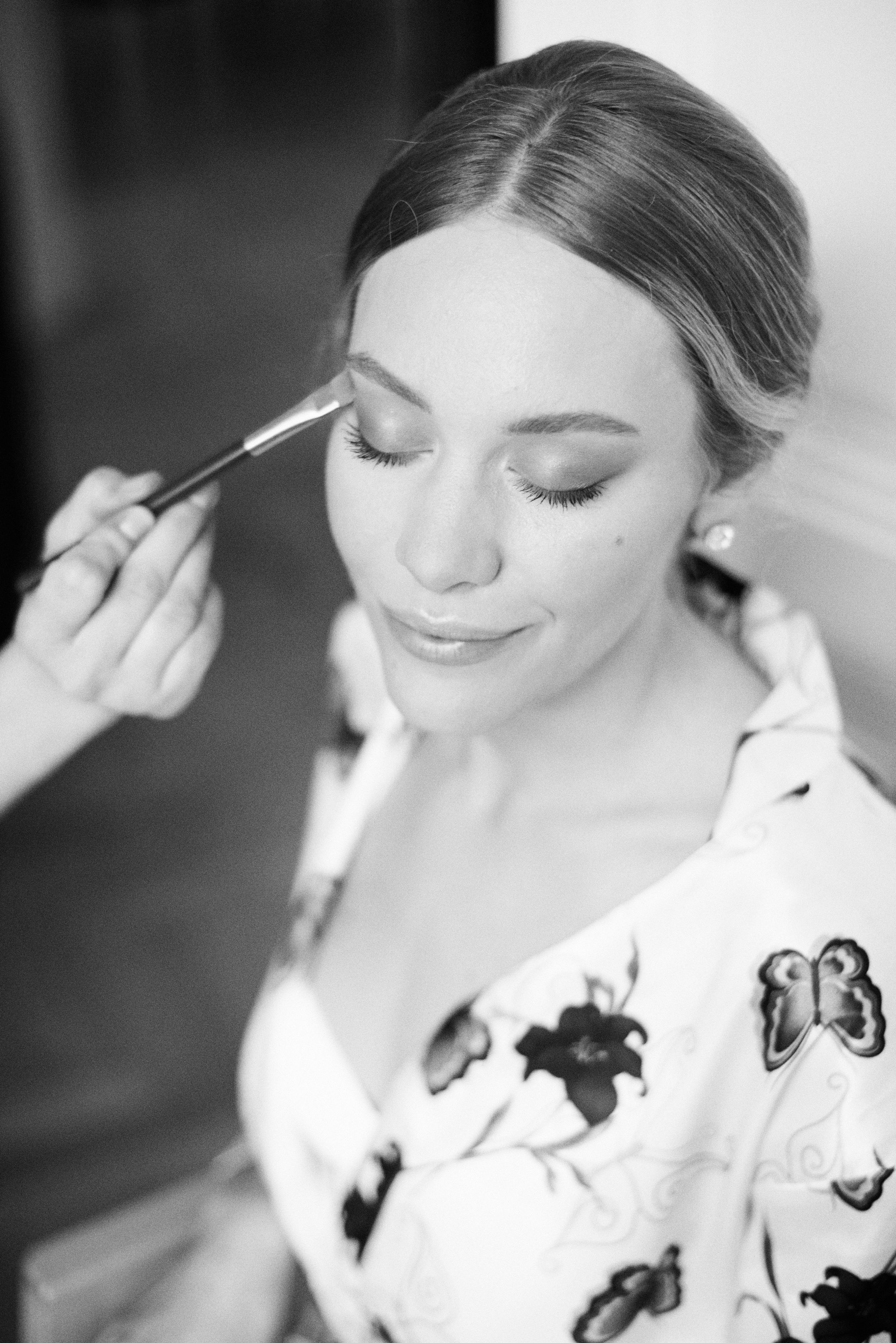 Black and white portrait of a bride having her makeup applied with a brush before her wedding.