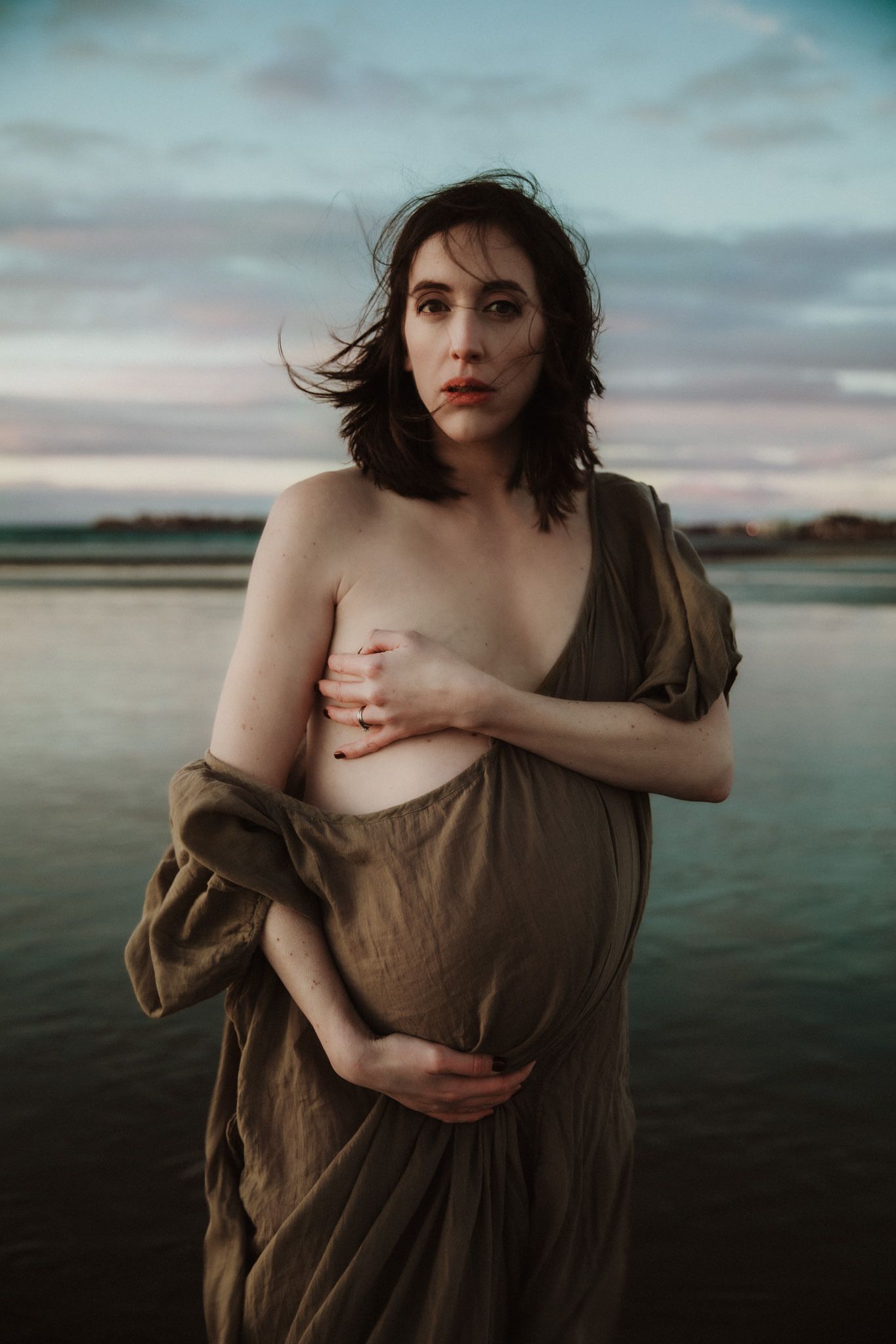 Pregnant woman standing in shallow water at sunset, holding her belly and looking towards the horizon.