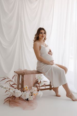 An elegant maternity photo featuring a woman in white seated on a bench with delicate floral decor.