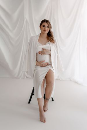 A minimalist maternity portrait featuring a woman in all white seated on a dark chair.