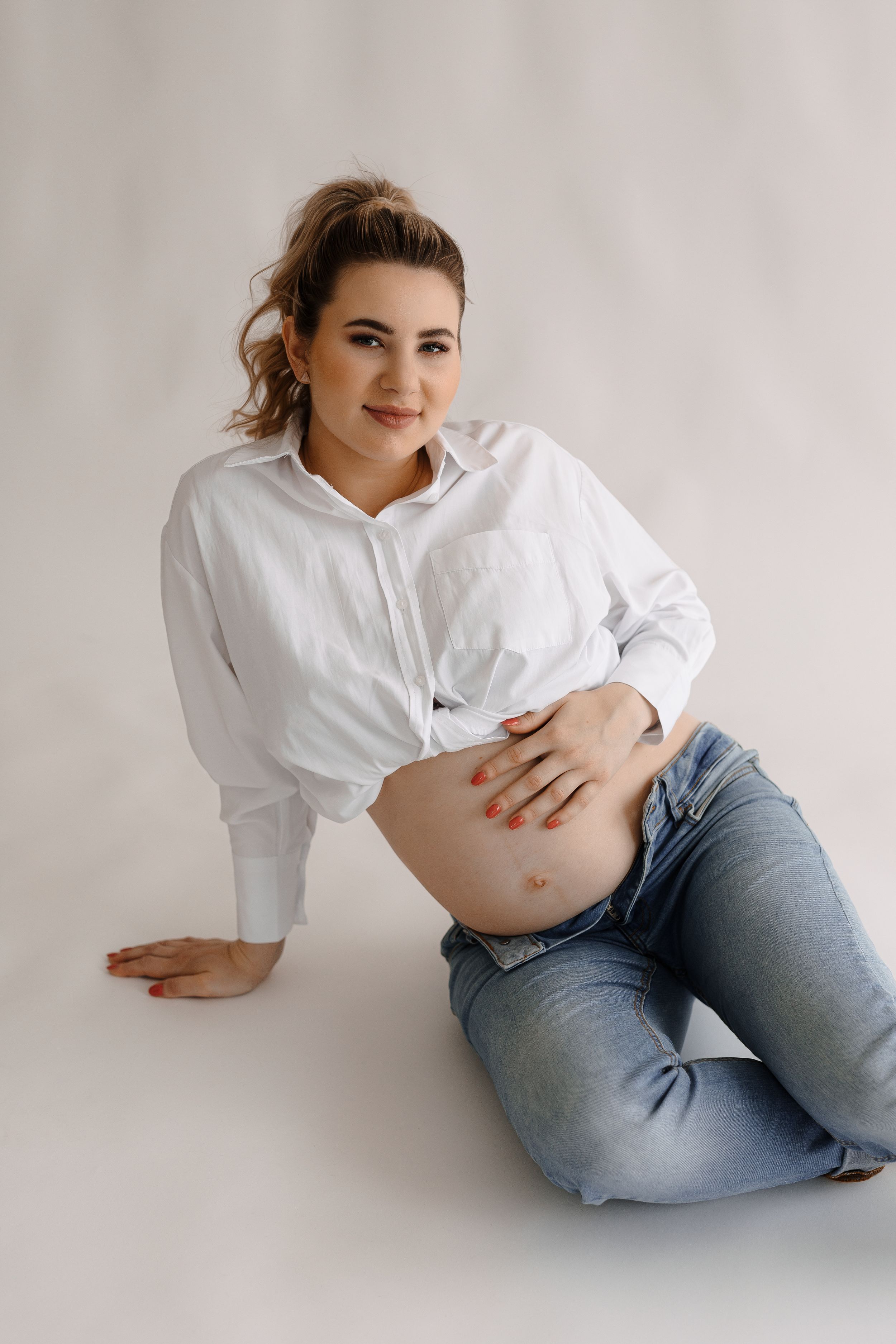 A casual maternity photo of a woman in jeans and white shirt against a neutral backdrop.