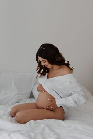 Intimate maternity portrait in white button-down shirt on white bedding showing pregnant belly in soft lighting.