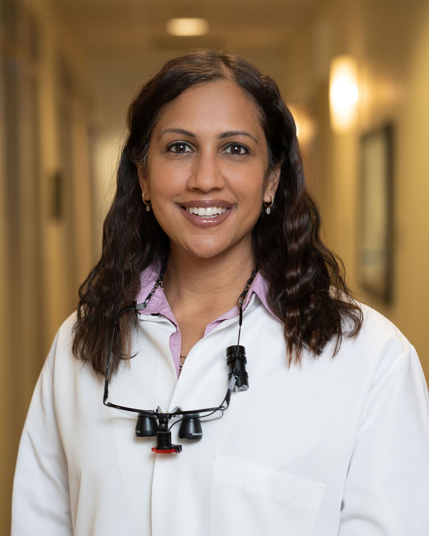 Smiling female dentist in white coat with loupes, standing in a medical hallway. Professional headshot by Digital Image House