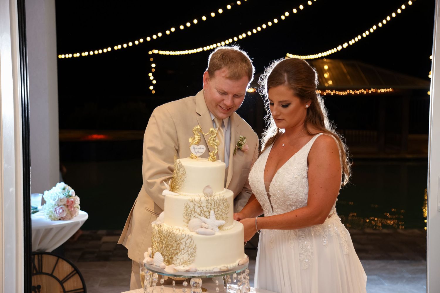 Newlyweds cut into their elegant white three-tiered wedding cake decorated with delicate details under string lights.