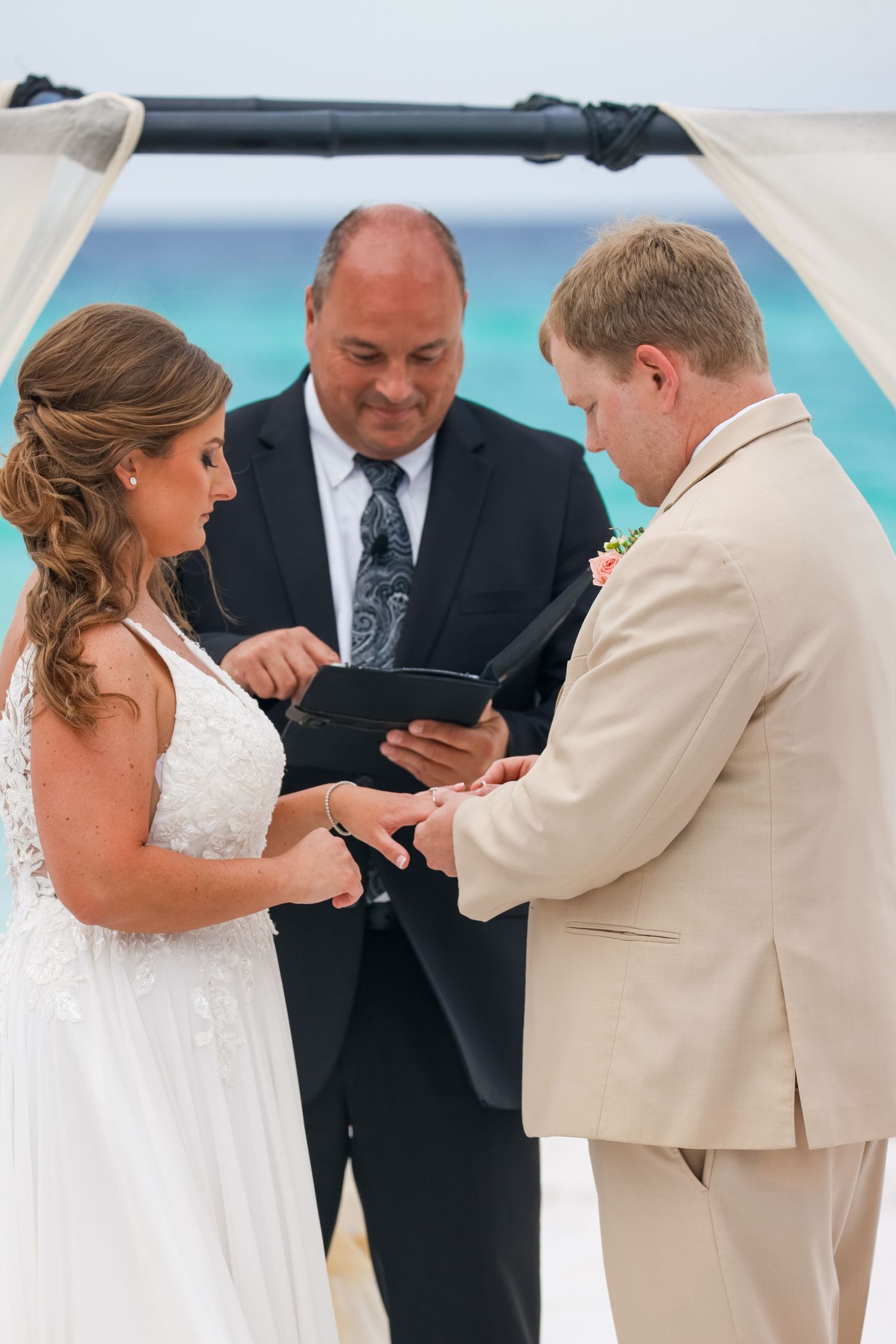 Wedding ceremony on beach with officiant and couple exchanging rings under white canopy.