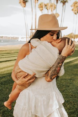 Person in hat and white dress holding baby against palm tree backdrop.