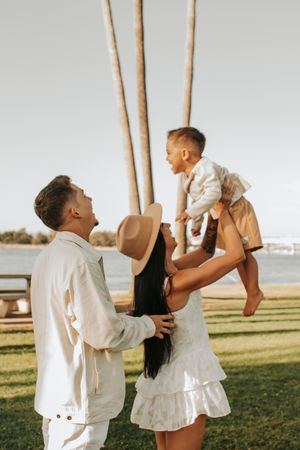 Family moment outdoors with parents lifting child against palm tree backdrop.