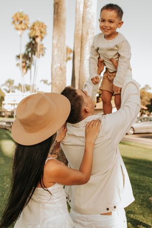 Family playfully lifting child in beige outfit with palm trees in background.