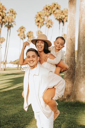 Happy family in white outfits playing and laughing outdoors with palm trees behind them.