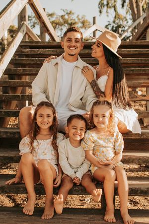 Family of five poses together on wooden stairs at sunset in coordinated neutral outfits.