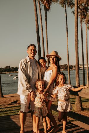 Family of five poses by waterfront with palm trees during sunset photoshoot.