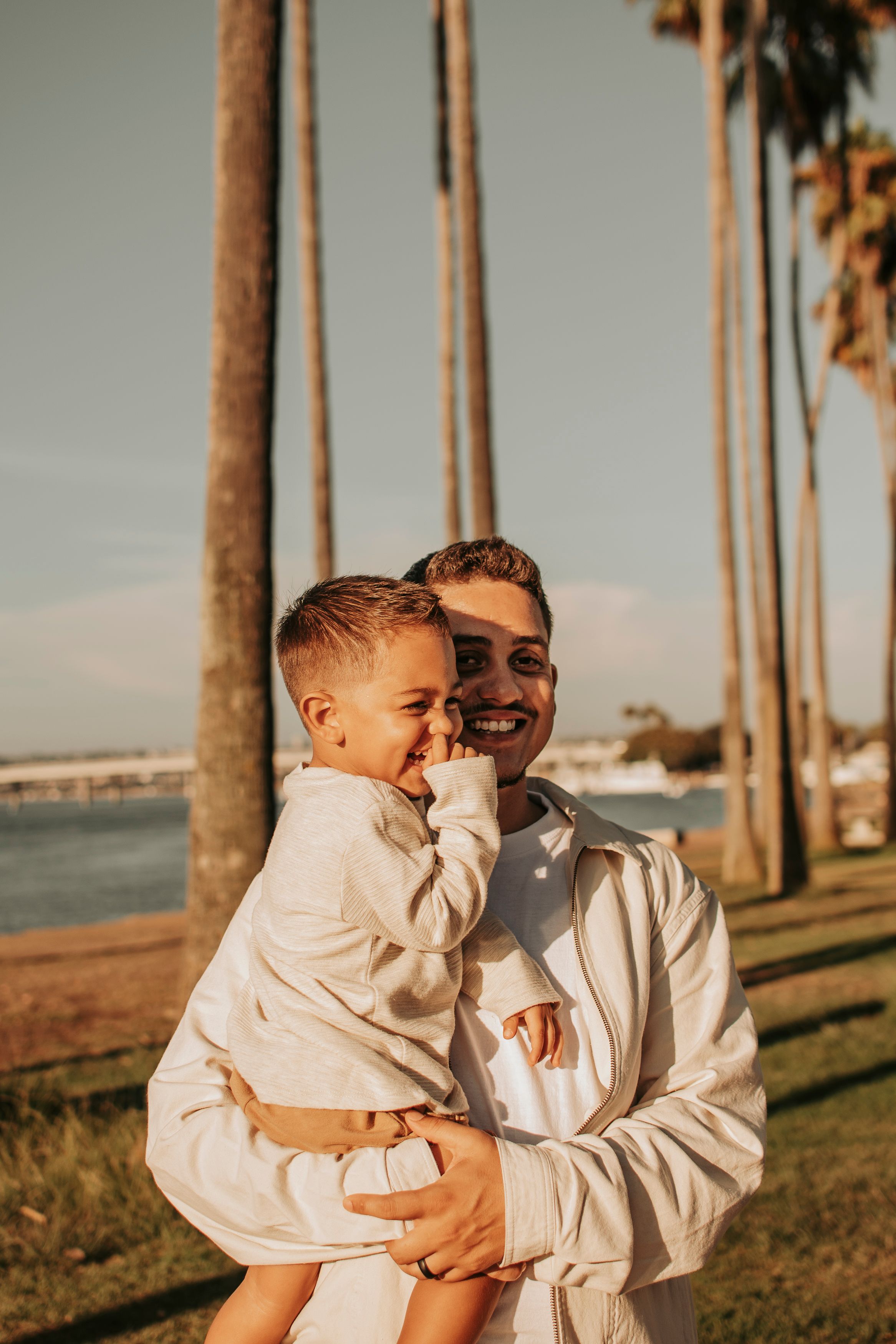 A heartwarming outdoor photoshoot featuring a baby in white and an adult by palm trees at sunset.