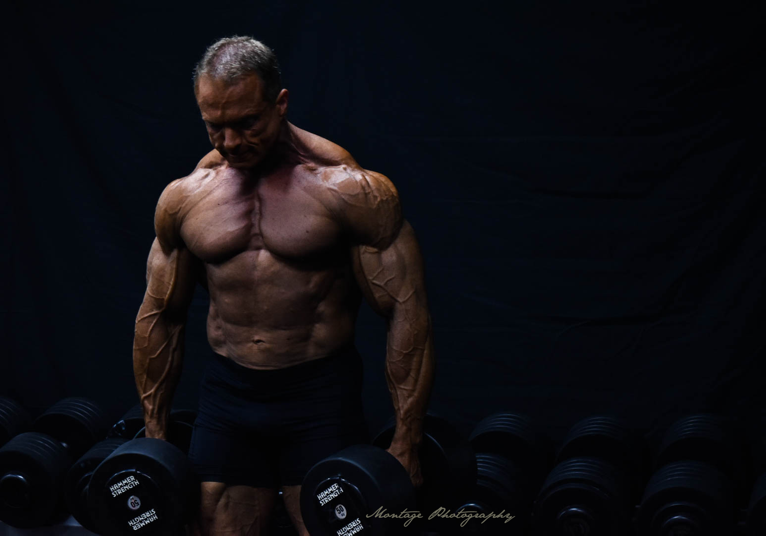 Dramatic low-key photo of bodybuilder training with dumbbells against dark background.