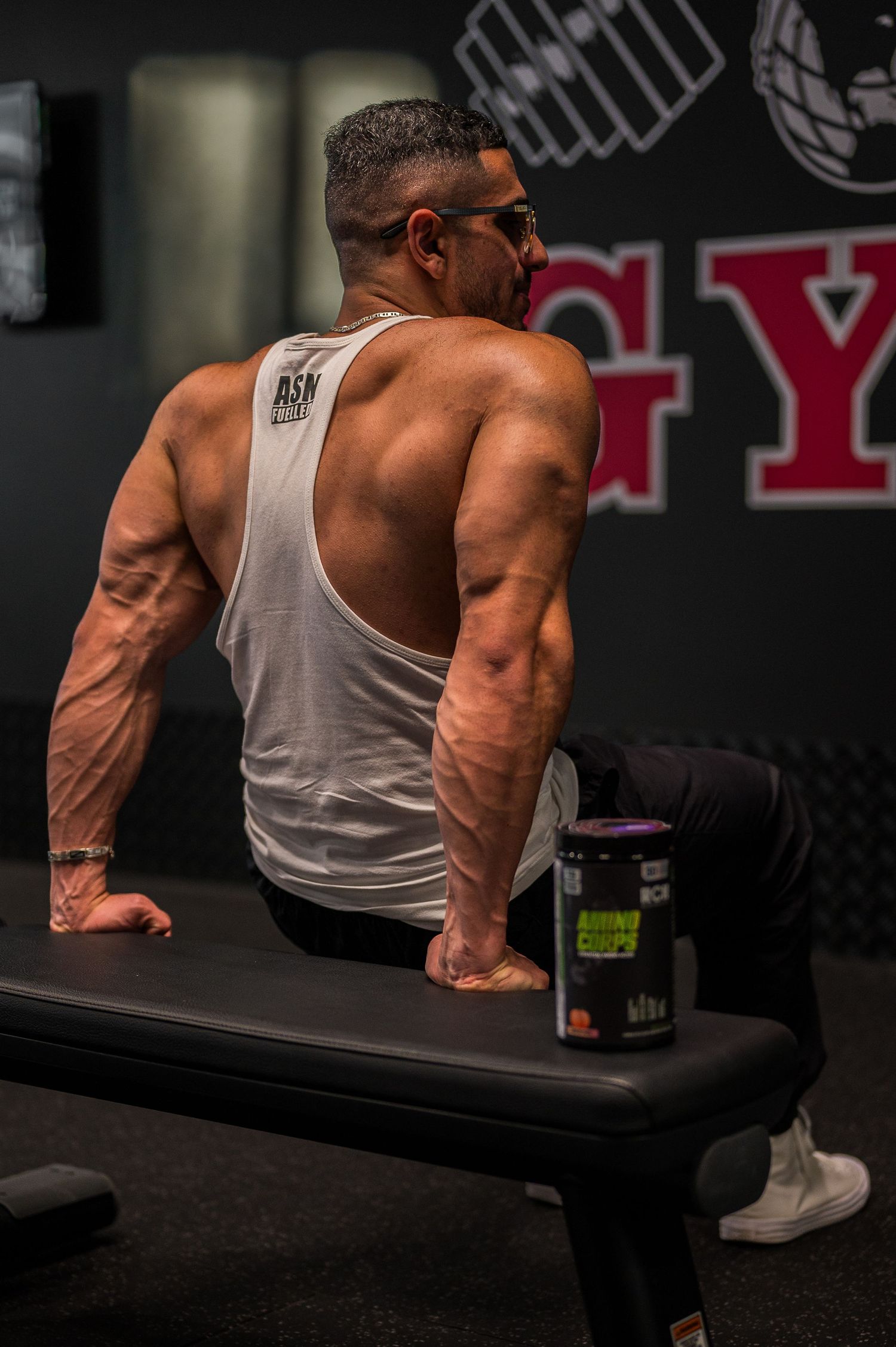 Athletic physique shown from behind in white tank top at gym with supplement container.