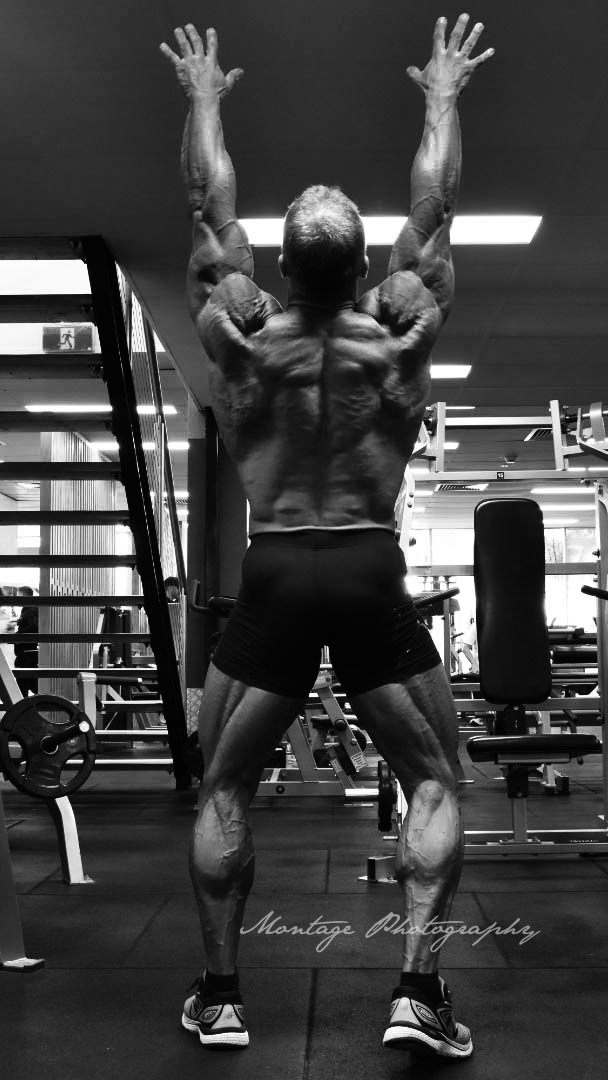 Muscular bodybuilder with defined back muscles stretches arms upward in black and white gym photo.