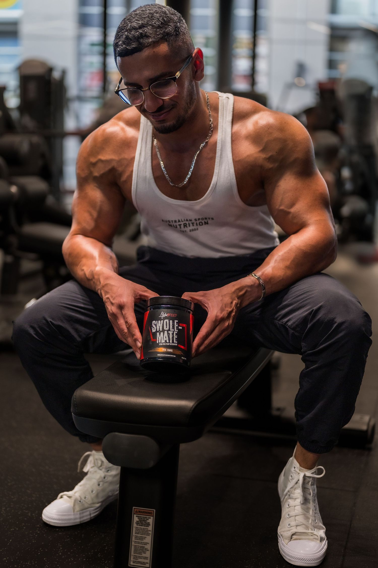 Muscular athlete in white tank top holding supplement container while sitting on gym bench.