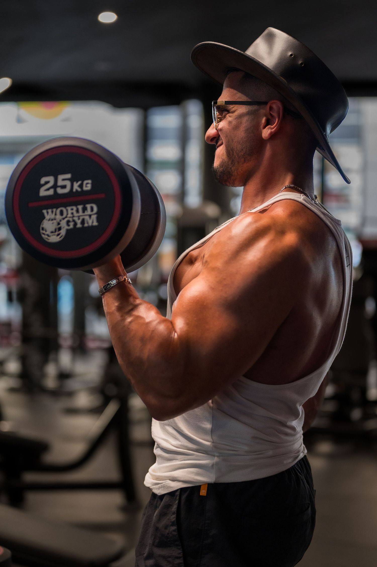 Athletic figure lifting 25kg dumbbell while wearing a cowboy hat in modern gym setting.